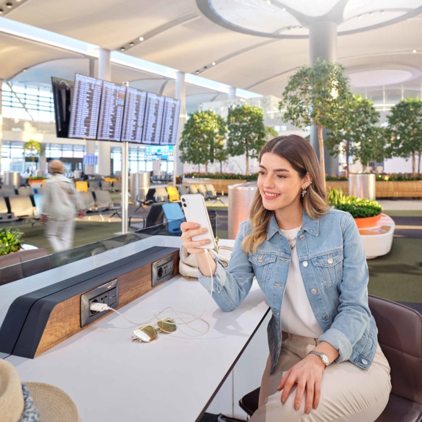 Eine junge Frau mit Smartphone sitzt in einem Wartebereich am Flughafen.