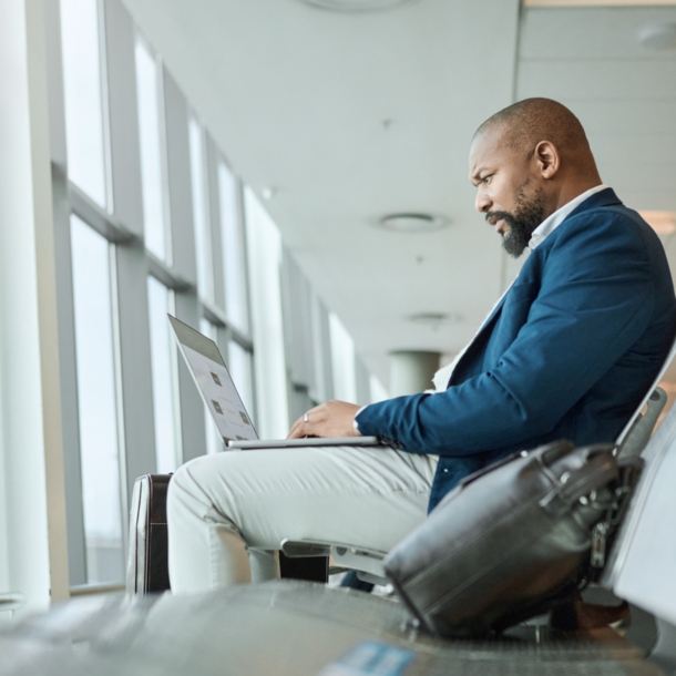 Ein Mann, der am Flughafen am Fenster sitzt und einen aufgeklappten Laptop auf dem Schoß hat.