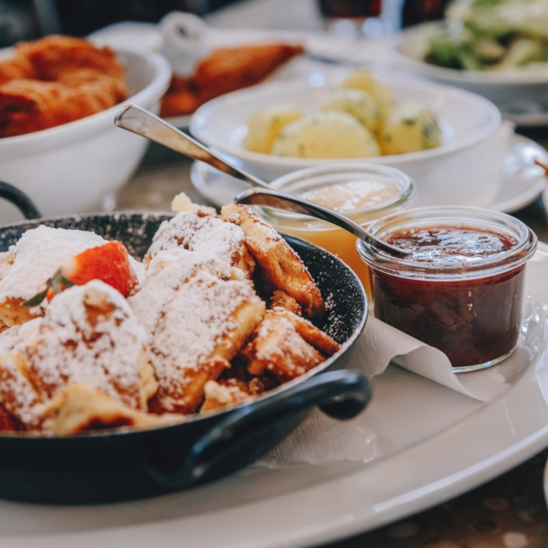 Ein schwarzer Topf mit Kaiserschmarrn auf einem weißen Teller, drumherum kleine Schälchen mit verschiedenen Leckereien.