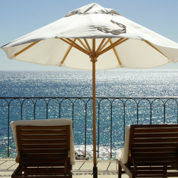 Terrasse und Geländer mit Blick aufs Meer, auf der mehrere Liegestühle und ein weißer Sonnenschirm stehen.