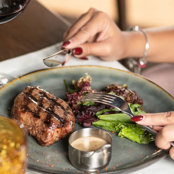 Nahaufnahme von zwei Händen mit Messer und Gabel über einem Stück Steak auf einem Teller an einem gedeckten Tisch.