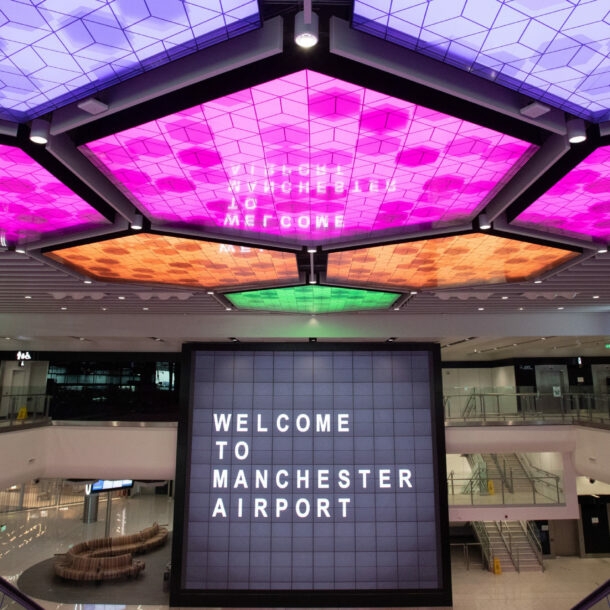 Menschenleere Flughafenhalle mit einem Bildschirm im Mittelpunkt, auf dem „Welcome to Manchester Airport“ steht und bunt erleuchteten LED-panels an der Decke.