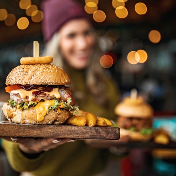 Ein Burger auf einem Brett, gehalten von einer Frau in Unschärfe in einem Lokal.