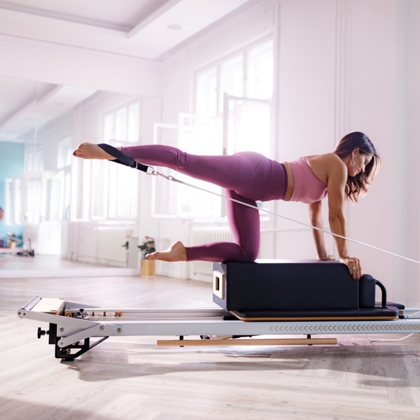 Eine Frau in pinker Sportkleidung kniet auf einer Box auf einem Pilates Reformer in einem hellen Raum.