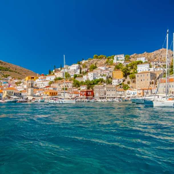 Mediterrane Ortschaft in Hügellage mit Hafenpromenade, im Vordergrund türkisblaues Meer mit Booten.