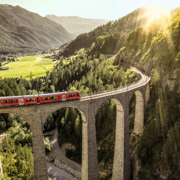 Ein roter Zug fährt auf einem Viadukt durch eine grüne Berglandschaft.