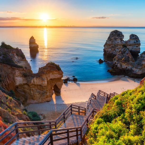 Der Strand Praia do Camilo in Portugal bei Sonnenuntergang, zu dem über Felsen eine Holztreppe nach unten führt.