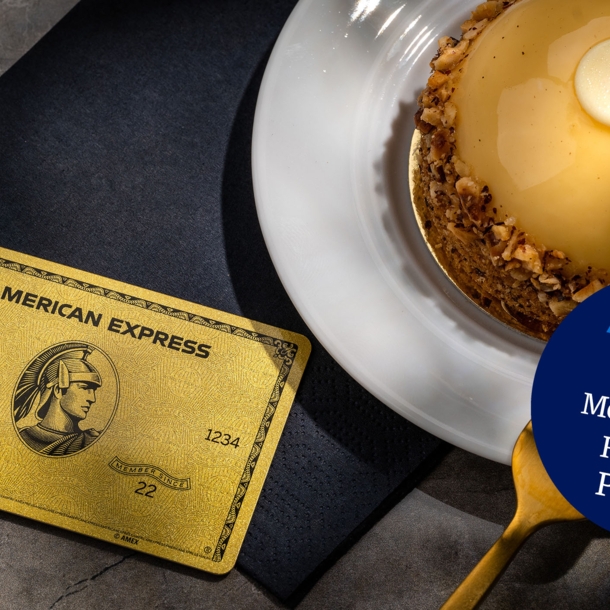 Eine goldene Kreditkarte von American Express liegt auf einer schwarzen Serviette neben einem goldfarbenen Törtchen auf einem Teller und einer goldenen Gabel.