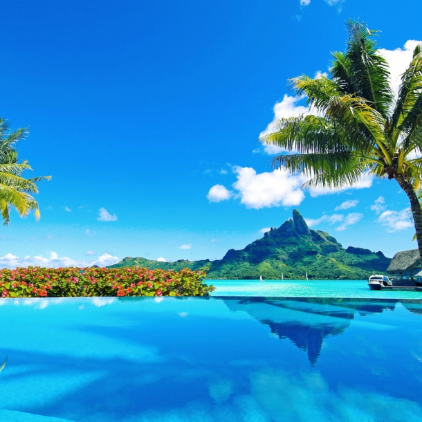 Von Palmen gesäumter Infinitypool an türkisblauer Lagune mit Blick auf eine Küste mit grüner Vegetation unter blauem Himmel.