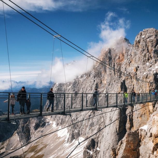 Personen auf einer Hängebrücke vor einer Felswand im Gebirge.