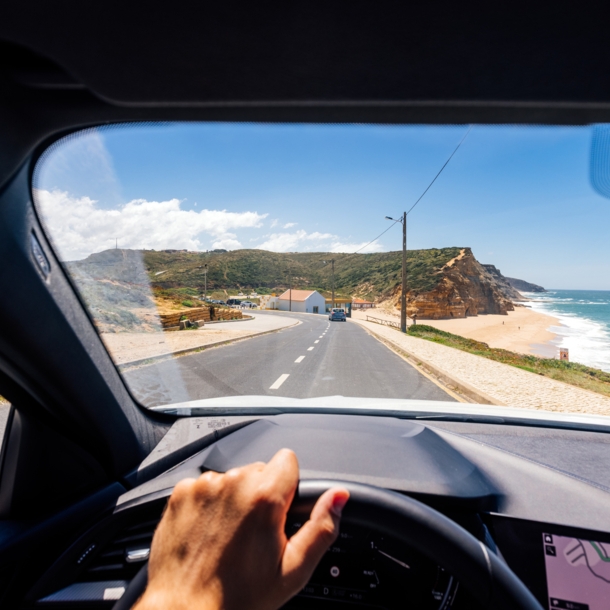 Blick über ein Lenkrad durch die Frontscheibe eines fahrenden Autos auf einer Straße an einer Küste mit Sandstrand bei Sonnenschein.