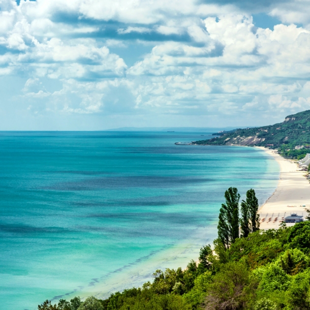 Grüner Küstenabschnitt mit Hotelanlagen an einem breiten Sandstrand vor flachem, türkisblauem Meer.