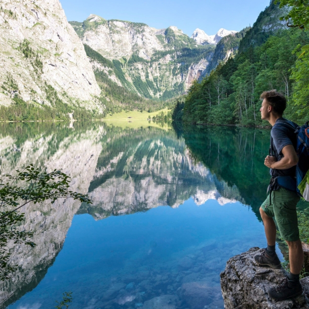 Eine Person mit Wanderrucksack steht auf einem Stein vor dem Königssee mit Blick auf die Berge.