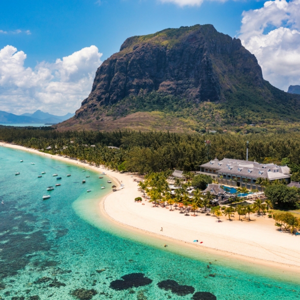 Küstenpanorama mit Hotelresort an einem palmengesäumten, weißen Sandstrand an türkisblauem Meer, im Hintergrund Berge.