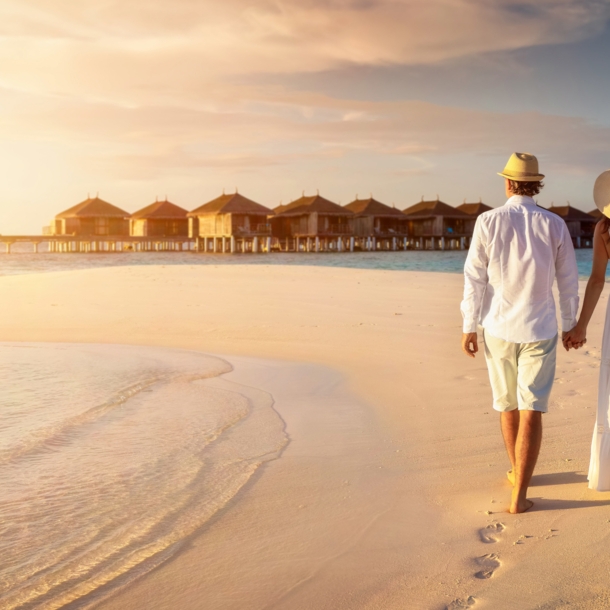 Rückansicht eines händchenhaltenden Paares in heller Sommerkleidung und mit Strohhüten an einem Strand vor einem Holzstegmit Wasserbungalows.