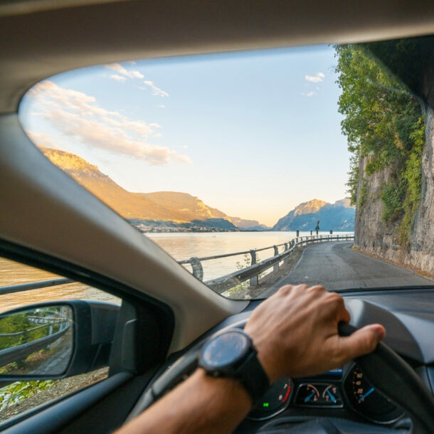 Blick auf eine Küstenstraße entlang einer Felswand durch die Windschutzscheibe eines Autos, im Vordergrund eine Hand am Lenkrad.