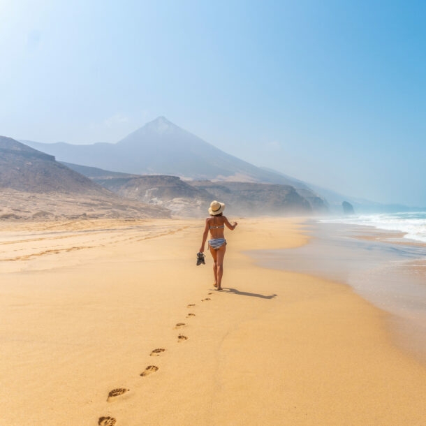 Rückansicht einer Frau im Bikini, die einen breiten Sandstrand entlangläuft, im Hintergrund spitze Felsformationen.