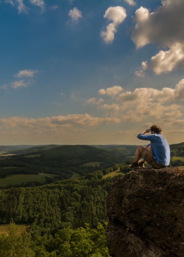 Ein Mann sitzt auf einem Felsen und blickt über die grüne Landschaft der Eifel