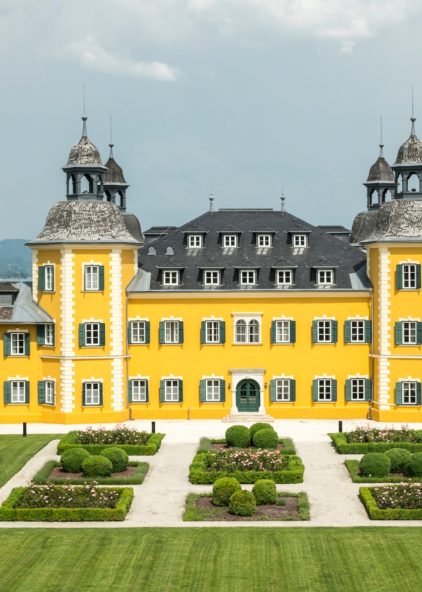 Ein gelbes Schlosshotel vor dem Panorama des Wörthersees