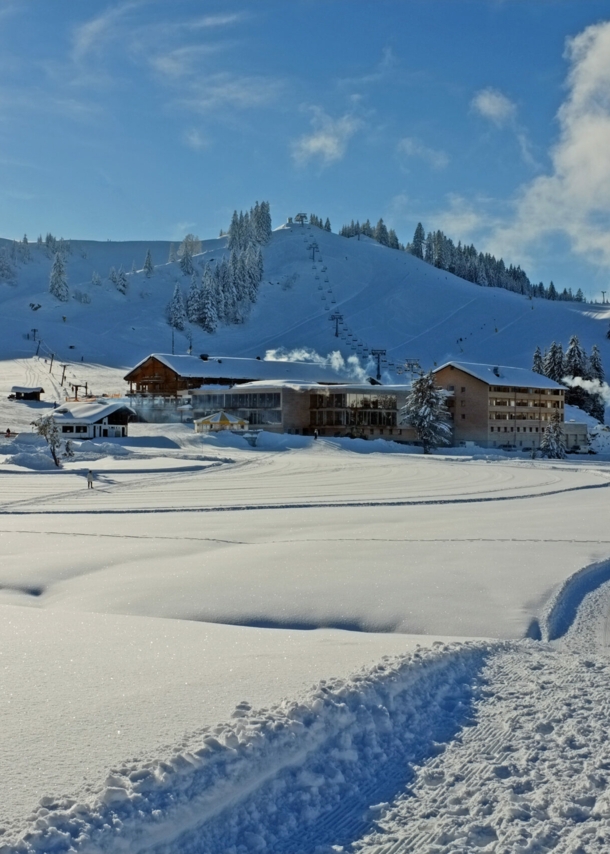 Loipen neben Neuschnee, im Hintergrund Hütten und ein schneebedeckter Berg.