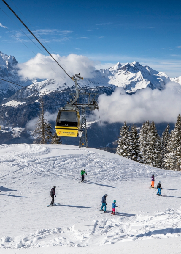 Skipiste mit Wintersportler:innen über die eine Seilbahn läuft, im Hintergrund Bergpanorama