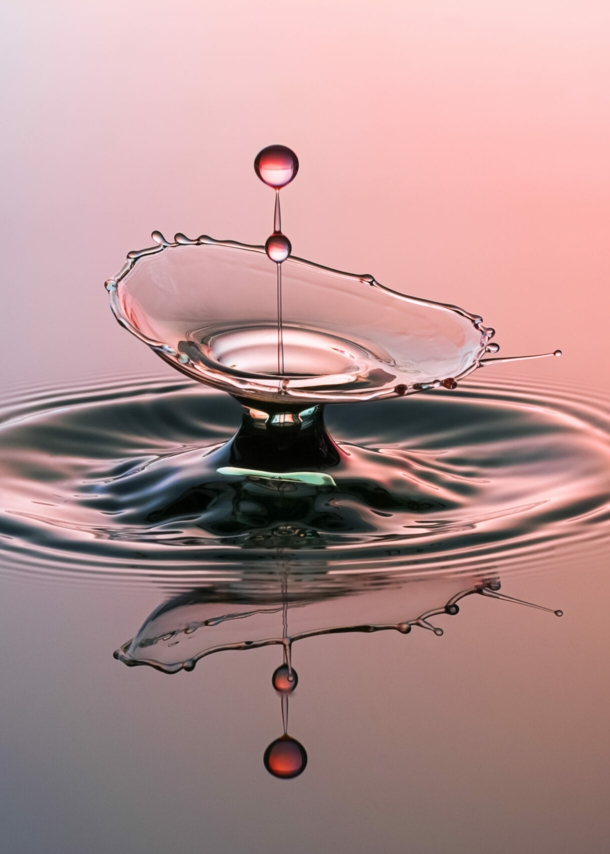 Eine lilafarben und rot angestrahlte Wasserfläche, in die ein Wassertropfen fällt.