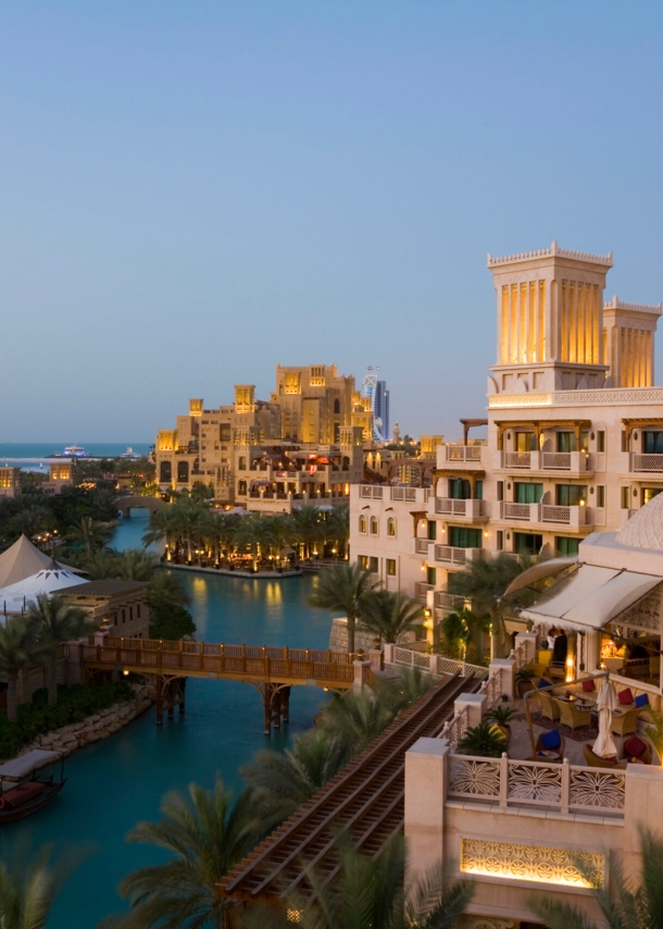 Stadtteil von Dubai mit Gebäuden an einem Fluss in der Abenddämmerung, im Hintergrund das Hotel Burj Al Arab in Segelform am Wasser