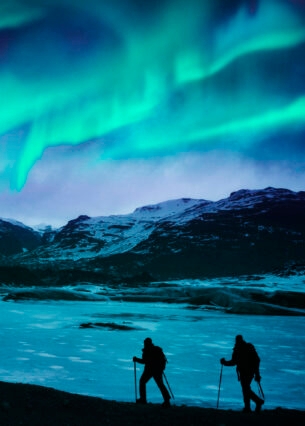 Zwei Wanderer vor einer Bergkulisse mit Polarlichtern.