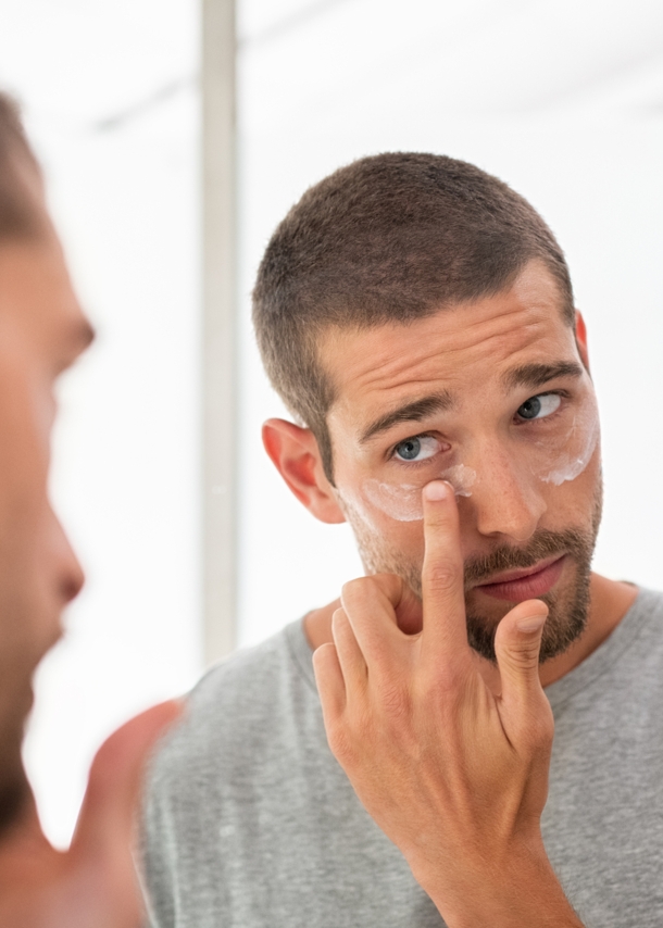 Ein Mann blickt in den Spiegel, während er sich unter den Augen Creme aufträgt