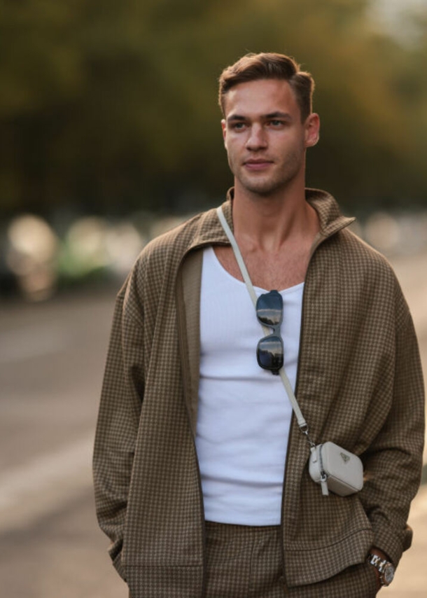 Ein Mann trägt einen braunen Trainingsanzug, hat eine Sonnenbrille am Shirt und eine kleine Tasche umgehängt.