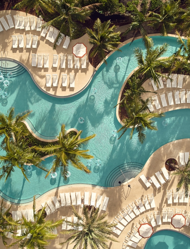 Blick von oben auf einen Pool in ungewöhnlicher Form mit Palmen und Liegen