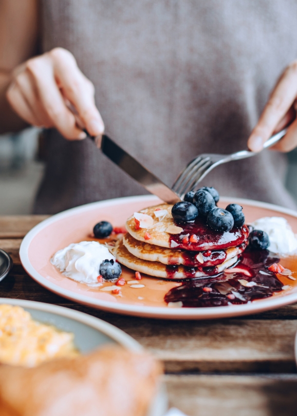 Eine Person sitzt an einem gedeckten Tisch vor einem Teller mit Pancakes im Außenbereich eines Cafés