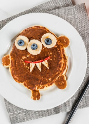 Frühstücksteller mit einem Pancake in Form eines kleinen Monsters, dessen Augen aus Bananen und Blaubeeren bestehen