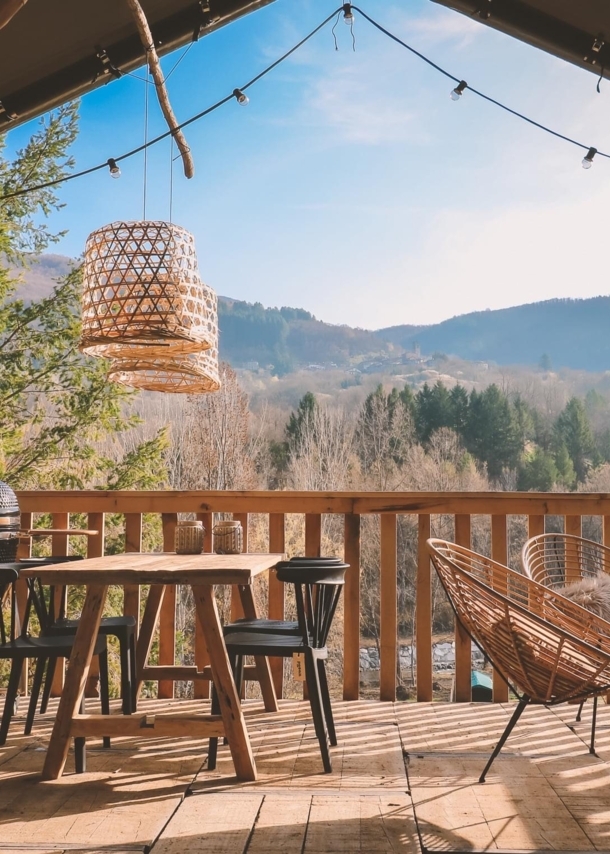 Holzterrasse mit Tisch, Stühlen und Lampen, im Hintergrund bewaldete Hügel