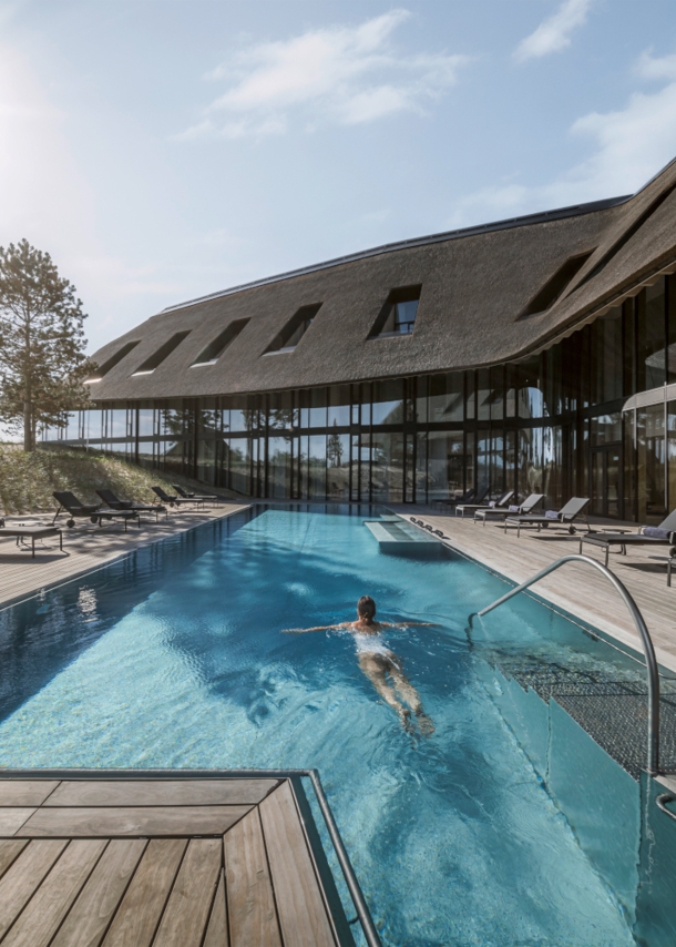 Ein luxuriöses Hotel in den Dünen mit Reetdach und Außenpool, in dem eine Frau schwimmt