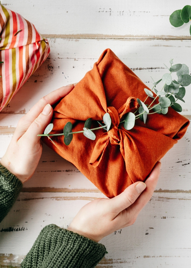 Ein Geschenk, verpackt in einem orangefarbenen Tuch und geschmückt mit einem grünen Zweig, wird von zwei Händen gehalten.