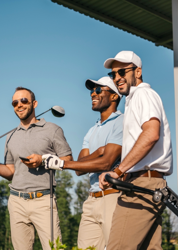 Drei Männer in Poloshirts, mit Sonnenbrillen und Golfschlägern stehen auf einer überdachten Terrasse und lachen