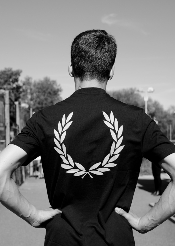 Rückansicht eines Mannes in einem dunklen T-Shirt mit Lorbeerkranz-Logo-Druck auf dem Rücken auf einem Tennisplatz, Schwarzweißaufnahme