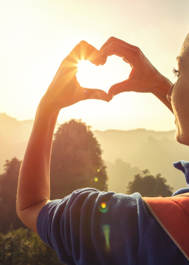 Eine junge Frau blickt in die Natur und formt ihre Hände zu einem Herz, durch das die Sonne scheint