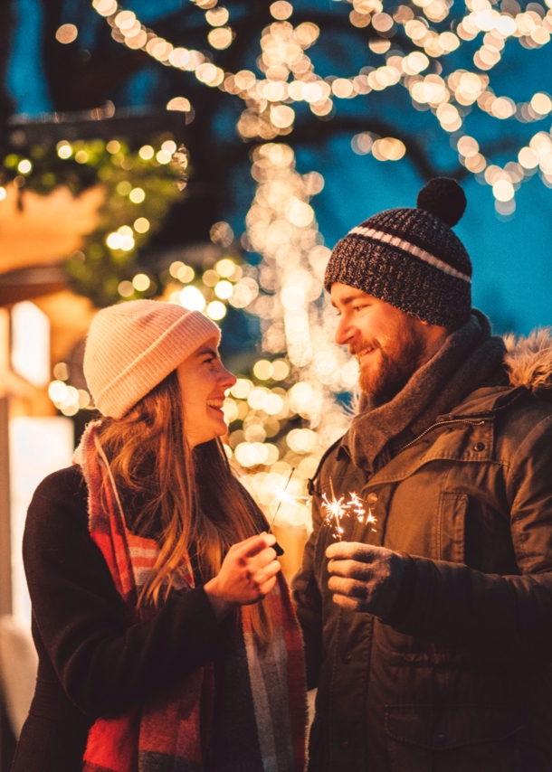 Eine Frau und ein Mann stehen auf dem Weihnachtsmarkt und schauen sich an, in ihren Händen halten sie Wunderkerzen