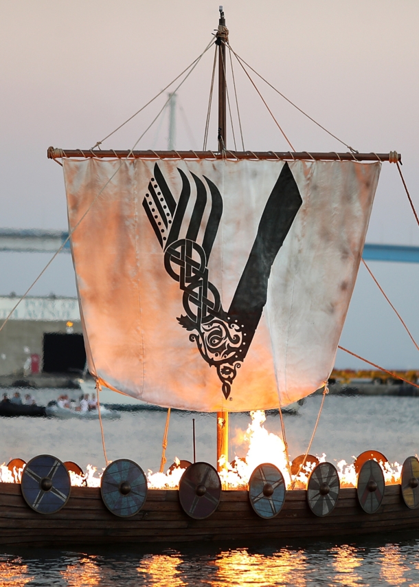 Aufnahme eines brennenden Wikingerschiffs auf dem Meer mit einem Segel, das das Logo der „Vikings“-Serie zeigt