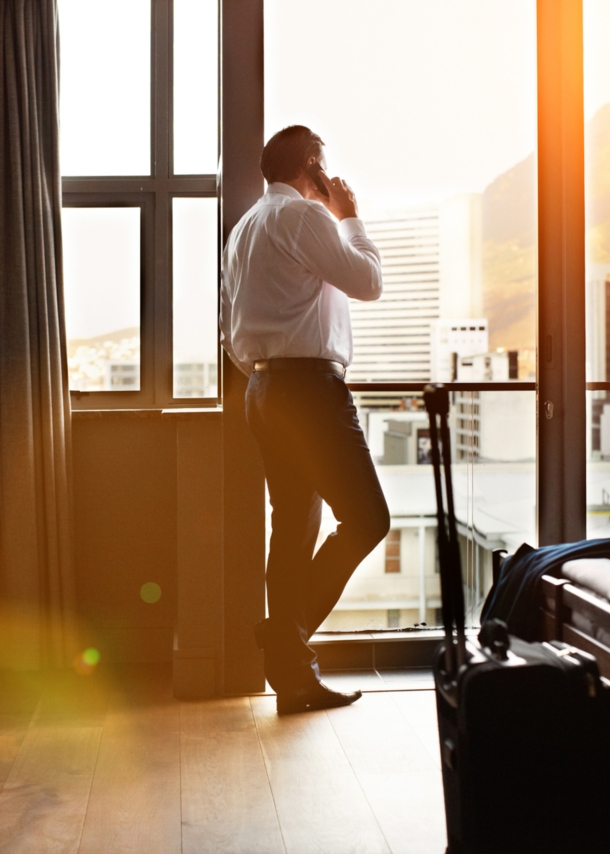 Rückansicht eines Geschäftsmannes, der durch ein Hotelzimmerfenster schaut und auf einem Mobiltelefon spricht