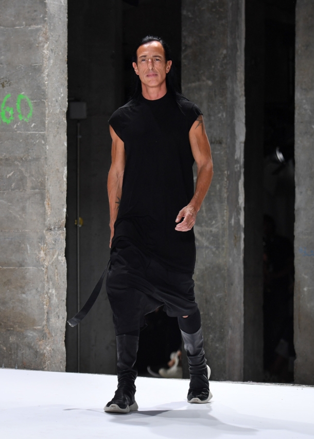 Designer Rick Owens in einem schwarzen, lässigen Kleid läuft in einer Industriehalle über einen Lauftsteg