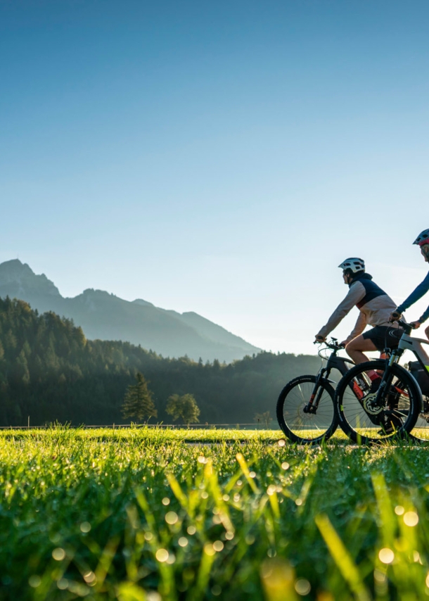 Zwei Personen fahren auf Mountainbikes durch eine Grasebene vor Bergpanorama im Sonnenschein