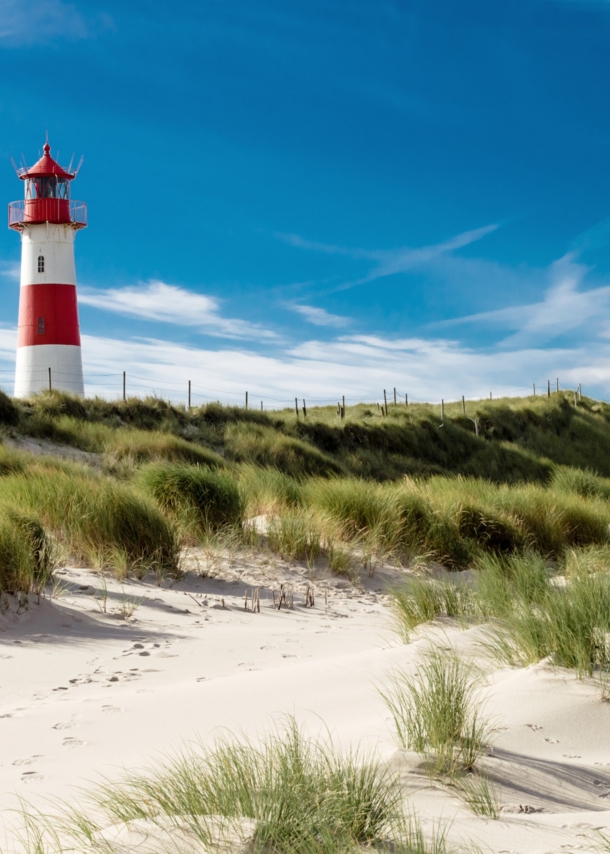 Ein rot-weiß geringelter Leuchtturm in einer grasbewachsenen Dünenlandschaft vor blauem Himmel