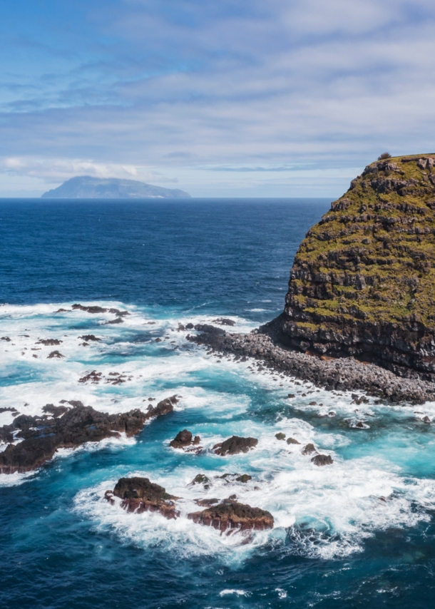 Luftaufnahme einer Steilklippe mit Leuchtturm und der Küste vorgelagerte Felsen im Meer, im Hintergrund eine Insel