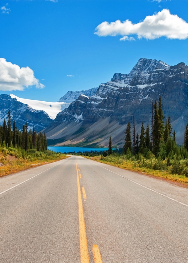 Ein Highway, umringt von Nadelbäumen, im Hintergrund schneebedeckte Berge und ein türkisblauer See.