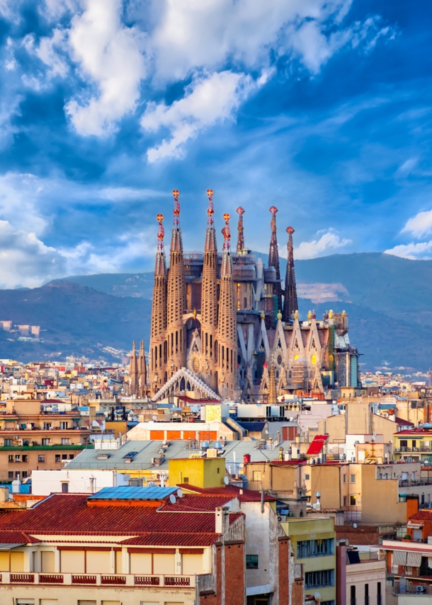 Stadtpanorama von Barcelona mit der emporragenden Sagrada Familia vor einem Gebirgszug im Hintergrund.