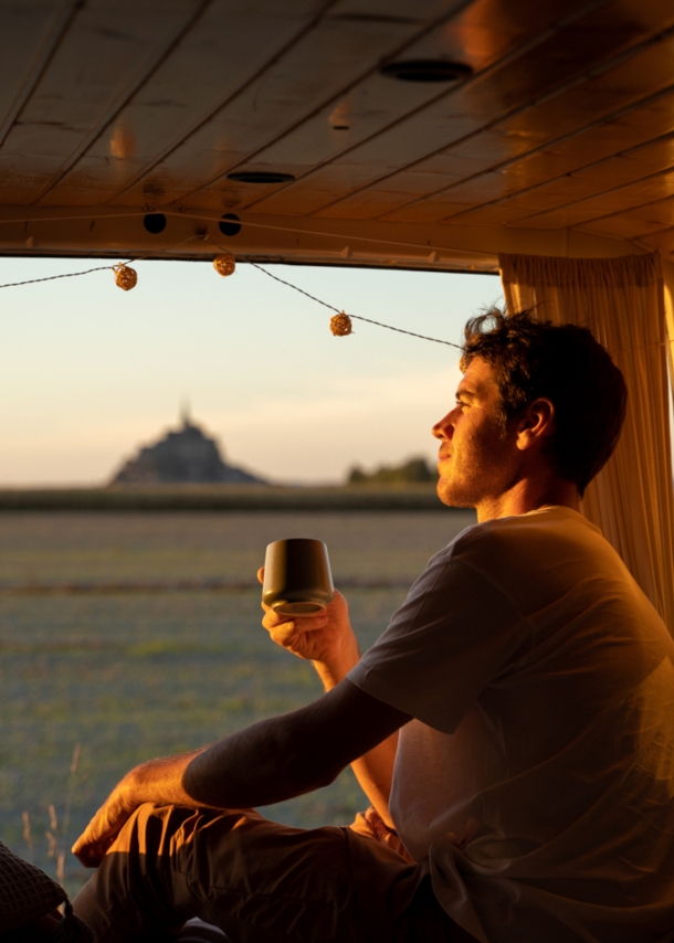 Ein junger Mann sitzt mit einem Kaffeebecher in der Hand im geöffneten Campervan im goldenen Sonnenlicht und blickt hinaus in die Natur.