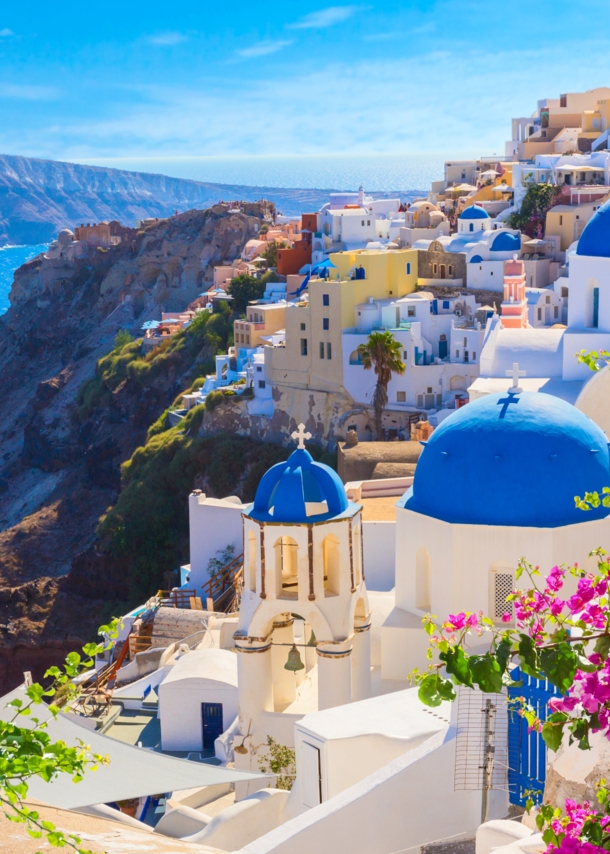 Steilküste von Santorin am Meer mit weißen Häusern mit blauen Dächern, im Vordergrund Bougainvillea.